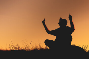 the silhouette of a man sitting outside on the grass and raising two hands up