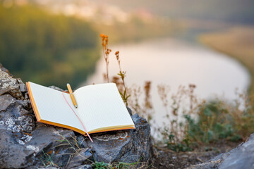 an open paper diary is located on a large stone next to the grass on a cliff, the background is blurred outside the forest and the river