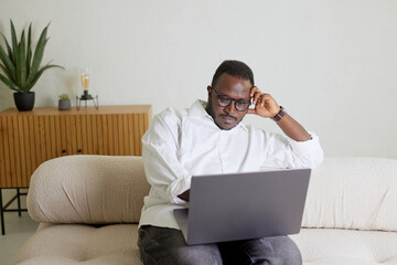 Handsome Black African American Man Working on Laptop Computer while Sitting on a Sofa in Cozy Living Room. Freelancer Working From Home. Browsing Internet, Using Social Networks, Having Fun in Flat.