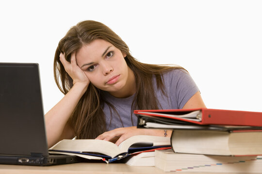 Young woman sitting in front of laptop beside a pile of thick textbooks while reading one with a frustrated stressed expression
