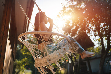 happy young female riding on macrame swing chair near country house outdoors