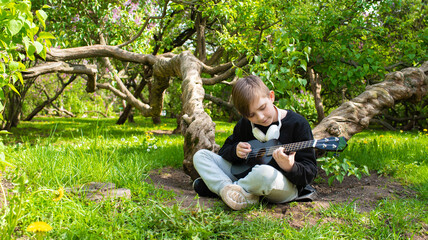 a little boy plays a small black ukulele, a guitar in a blooming garden, a park on a sunny day	