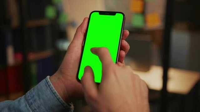 Shooting of male hands in jeans jacket holding smartphone with green screen. Man scrolling social media, reading message on gadget. Chroma key. Indoors. Blurred background of living room. Evening