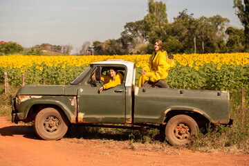 Duas apicultoras em uma caminhonete, parada em uma estrada de chão, visitando uma plantação de girassóis.
