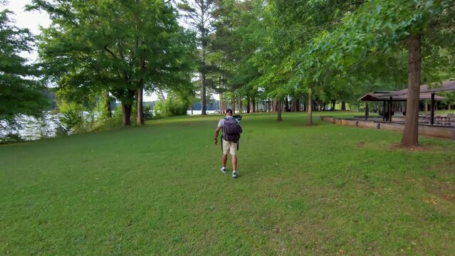 footage of an African American man wearing a backpack, holding a camera hiking in a park with lush green trees and grass at sunset at Proctor Landing Park at lake Acworth in Acworth Georgia USA