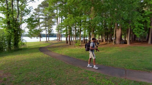 footage of an African American man wearing a backpack, holding a camera hiking in a park with lush green trees and grass at sunset at Proctor Landing Park at lake Acworth in Acworth Georgia USA