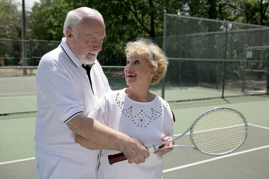 A senior couple playing tennis together.  He's showing her how to grip her racquet.