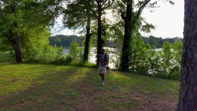 footage of an African American man wearing a backpack, holding a camera hiking in a park with lush green trees and grass at sunset at Proctor Landing Park at lake Acworth in Acworth Georgia USA