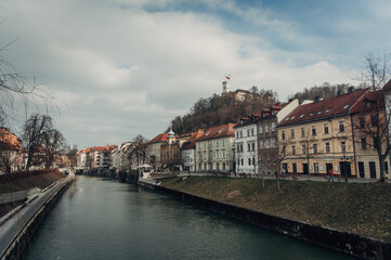 Fototapeta na wymiar This photo captures the Ljubljana river, historical buildings, and castle during a cloudy day at the end of winter, showcasing the beauty and charm of the Slovenian capital.