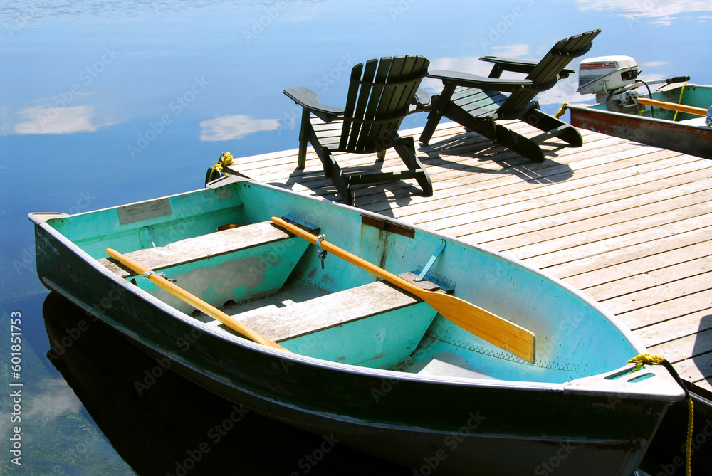 Wall mural Paddle boat and two adirondack wooden chairs on dock facing a blue lake