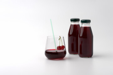 Two small bottles under caps and a glass with a cherry drink on a white background. In the glass there is a cherry straw, on the edge of the glass there is a cherry berry on the stalk.
