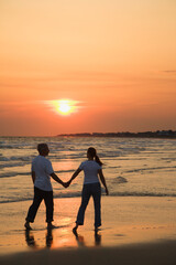 Mid-adult couple holding hands and walking on beach at sunset.