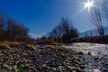 Cobble stones rolling on a wide river landscape on a winter leafless forest under a blue sunny sky with sun shining flare