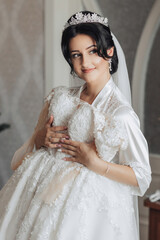 Portrait. A brunette bride in a robe and a chic crown is standing, holding her wedding dress and looking to the side. Gorgeous make-up and hair. Voluminous veil. Wedding photo. Beautiful bride