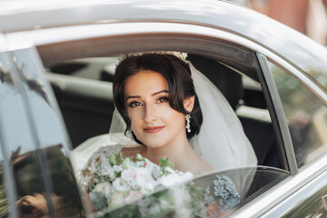 A brunette bride, smiling sincerely, sits in a black car on her wedding day with a bouquet of white roses. Portrait of the bride. Beautiful curls. Beautiful makeup and hair. A luxurious white dress