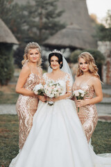 Obraz na płótnie Canvas A brunette bride in a white elegant dress with a crown and her blonde friends in gold dresses pose with bouquets. Wedding portrait in nature, wedding photo in a light tone.