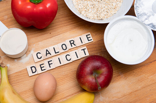 Inscription calorie deficit next to fruit, eggs, protein powder and oatmeal. Weight reduction. Healthy sources of proteins and carbohydrates