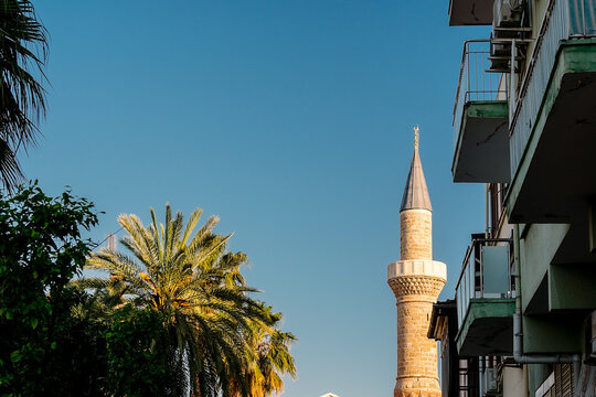 Minaret of the Tekeli Mehmet Pasha Mosque in the Turkish city of Antalya. Sunlit minaret between buildings and trees.