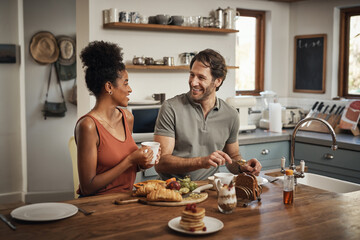 Happy interracial couple, breakfast and morning in kitchen for healthy meal, talk or bonding at home. Man and woman smiling for coffee, conversation or tea and food on weekend together by the table