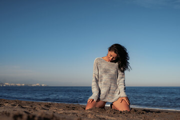 Fototapeta na wymiar beautiful brunette woman in a sweater and swimsuit sits on the sand by the sea enjoys the environment