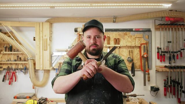 young carpenter with a protective apron holding a wooden mallet and a chisel with his hands crossed. High quality 4k footage