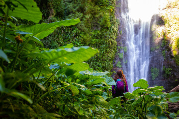 back view of adventurous hiker girl standing in front of large tropical waterfall surrounded by...