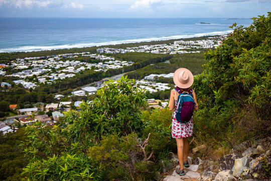 beautiful girl in a hat walking down the trail from mount coolum with panorama of sunshine coast and coolum beach in the background; hiking in queensland, australia