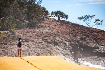 lonely woman walking on the sand dunes towards large cliff made of red rocks; walking alone on australian beach near agnes water in queensland, australia