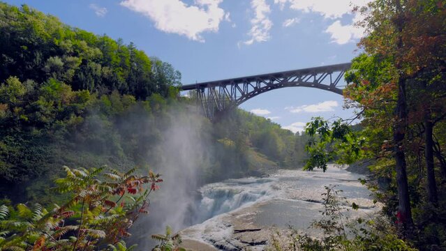 Tilt Down Letchworth State Park with Falls and Bridge Slow Framed Foliage. Wide moving down shot of the upper falls at Letchworth State Park under a bridge framed by foliage in slow motion