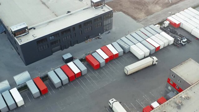 Aerial view of semi truck with cargo trailer moving along a warehouse with many containers standing on ramps for unloading and loading goods in a logistics park