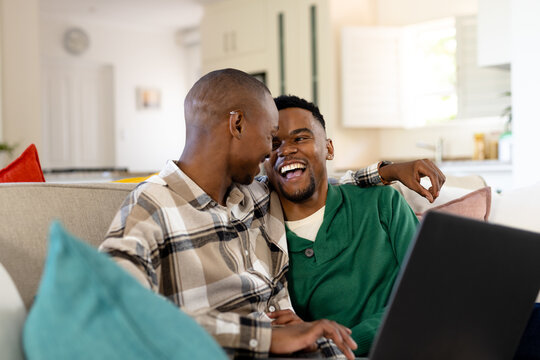 African american romantic gay couple looking at each other while watching movie over laptop on sofa