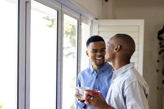 African american gay couple dressed in formals holding coffee cups and talking by window