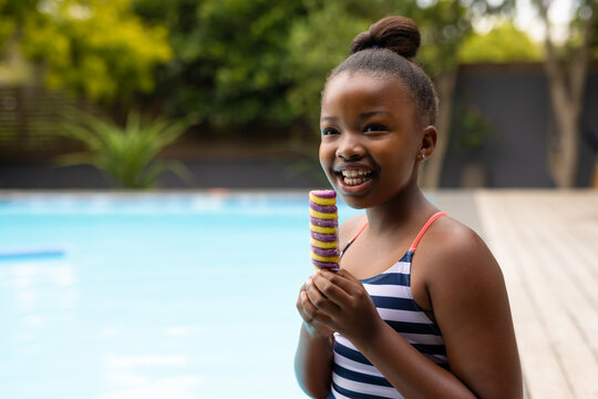 Happy african american girl in swimsuit sitting by pool eating ice cream, with copy space