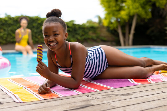 Happy african american daughter lying on towel by pool holding ice cream, mother in background