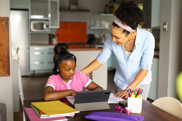 Happy african american mother helping daughter in headphones having online lesson on tablet at home