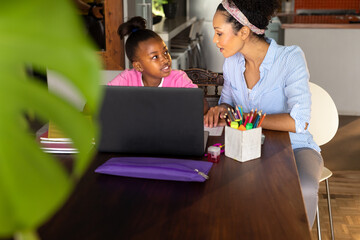 Smiling african american mother helping happy daughter doing schoolwork using laptop at home