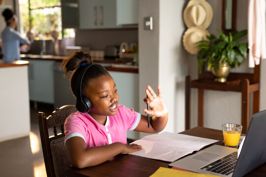 Happy african american daughter in headphones waving during online lesson using laptop at home