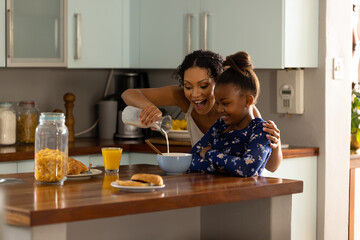 Happy african american mother preparing breakfast cereal for daughter sitting in kitchen island