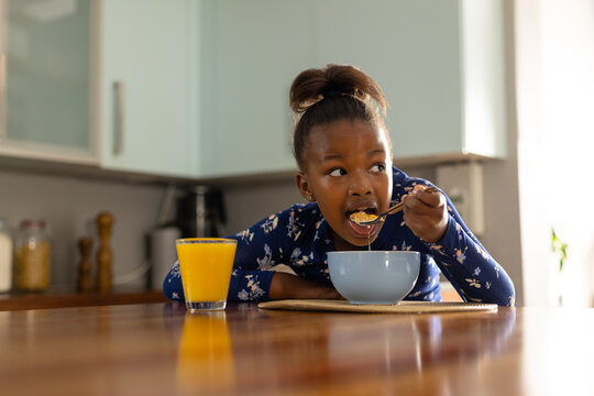 Happy african american girl eating breakfast cereal sitting in kitchen and looking away