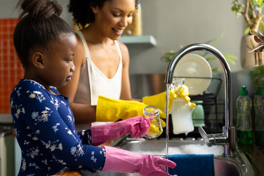 Happy african american mother and daughter washing up glasses and dishes standing at kitchen sink