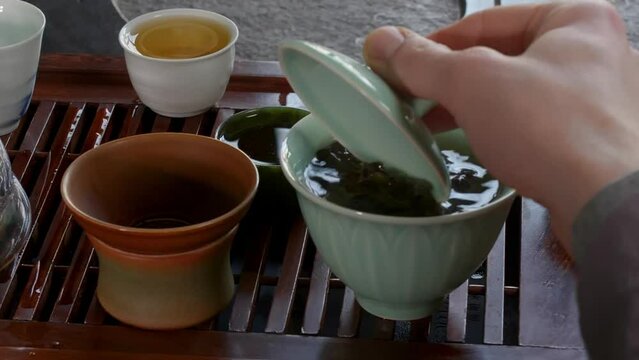 Tea master brewing fresh green tieguanyin tea in gaiwan bowl on wooden table chaban mixing leaves and water with lid, hands close-up. Professional traditional chinese tea ceremony preparing hot drink.