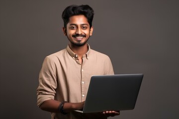 Handsome happy young Indian man holding laptop against a studio background. generative AI