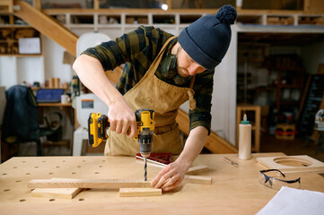 Young man jointer working with drill leaning over table at carpentry workshop - obrazy, fototapety, plakaty
