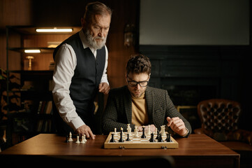 Senior man teaching young guy playing chess explaining rules of game