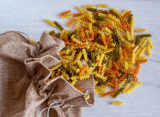 Uncooked multicolored Fusilli pasta spilled out of fabric bag over white wood background. top view.