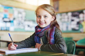 Education, portrait of young girl in classroom and writing in her notebook at her desk in school building. Studying or homework, learn or academic and happy female student with pen sitting on chair
