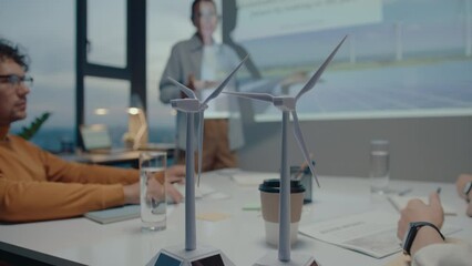Wind turbine models on meeting table in the office of eco-friendly company, colleagues discussing presentation in the background. Close-up shot
