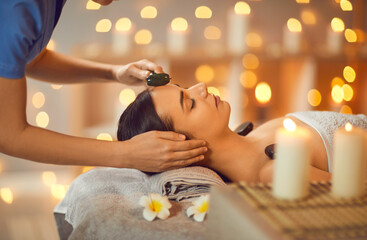 Obraz na płótnie Canvas Pretty woman is getting face massage at spa salon. Cosmetologist is doing facial massage with jade roller to relaxed young woman lying on towel with flowers on massage bed. Beauty, skin care concept