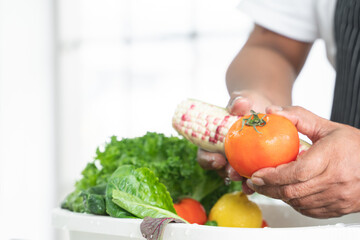 Close-up on hands of senior man washing tomato and corn at home. Vegetables, lettuce and lemon soak with water in basin or sink. Healthy food and cleaning before cooking concept. Focus on tomato