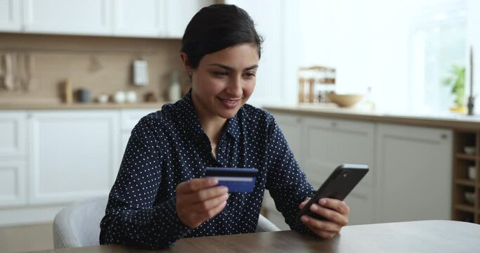 Smiling young Indian woman sit at home in kitchen, hold smart phone and credit card, enter personal data, makes purchase distantly, pay for e-service use modern application. Ecommerce retail shopping